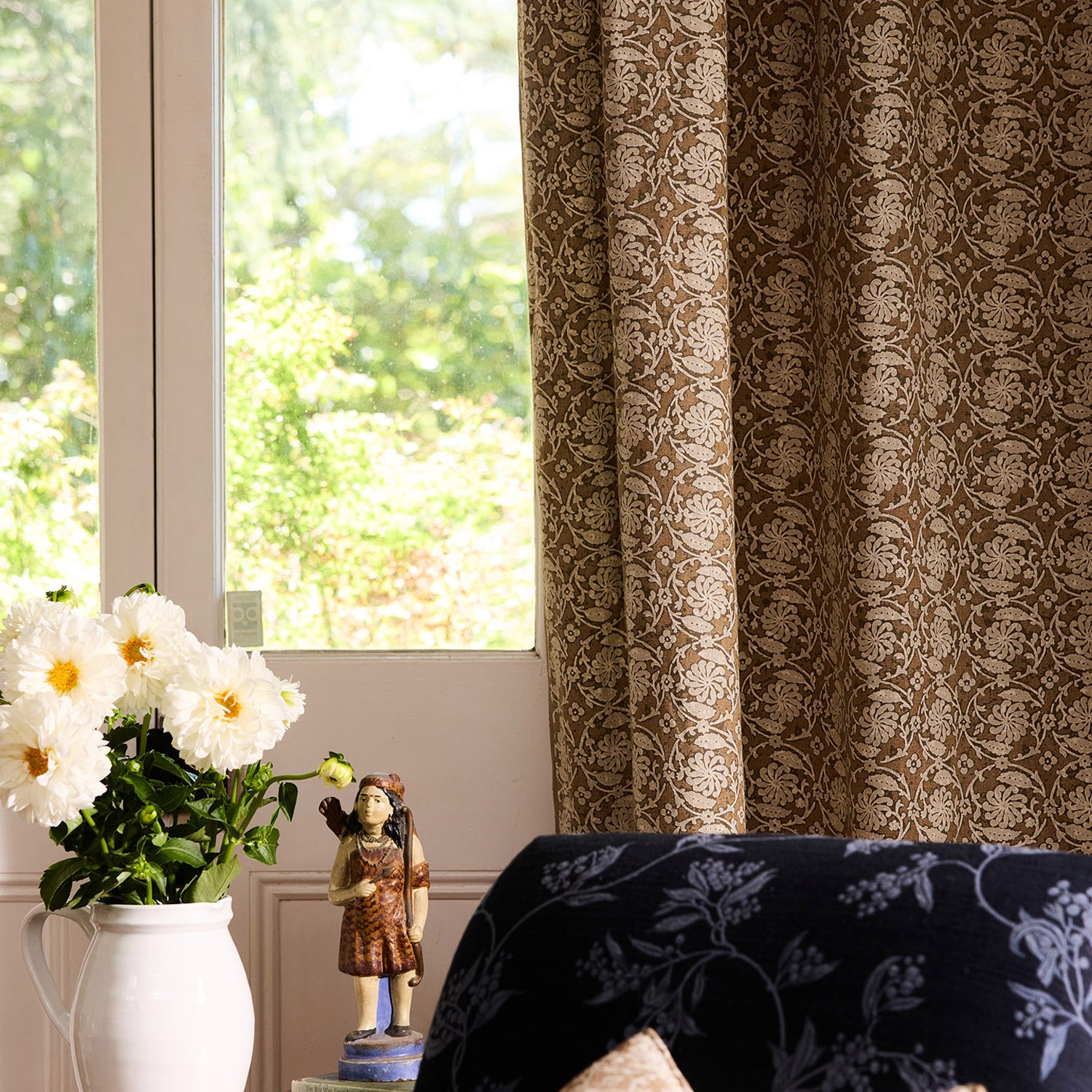 Decorative curtain with floral pattern in a room with a window and a vase of flowers on a table.