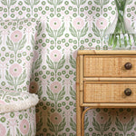 Upholstered side chair and wallpaper in matching pink and green floral pattern along side a rattan table with drawers.