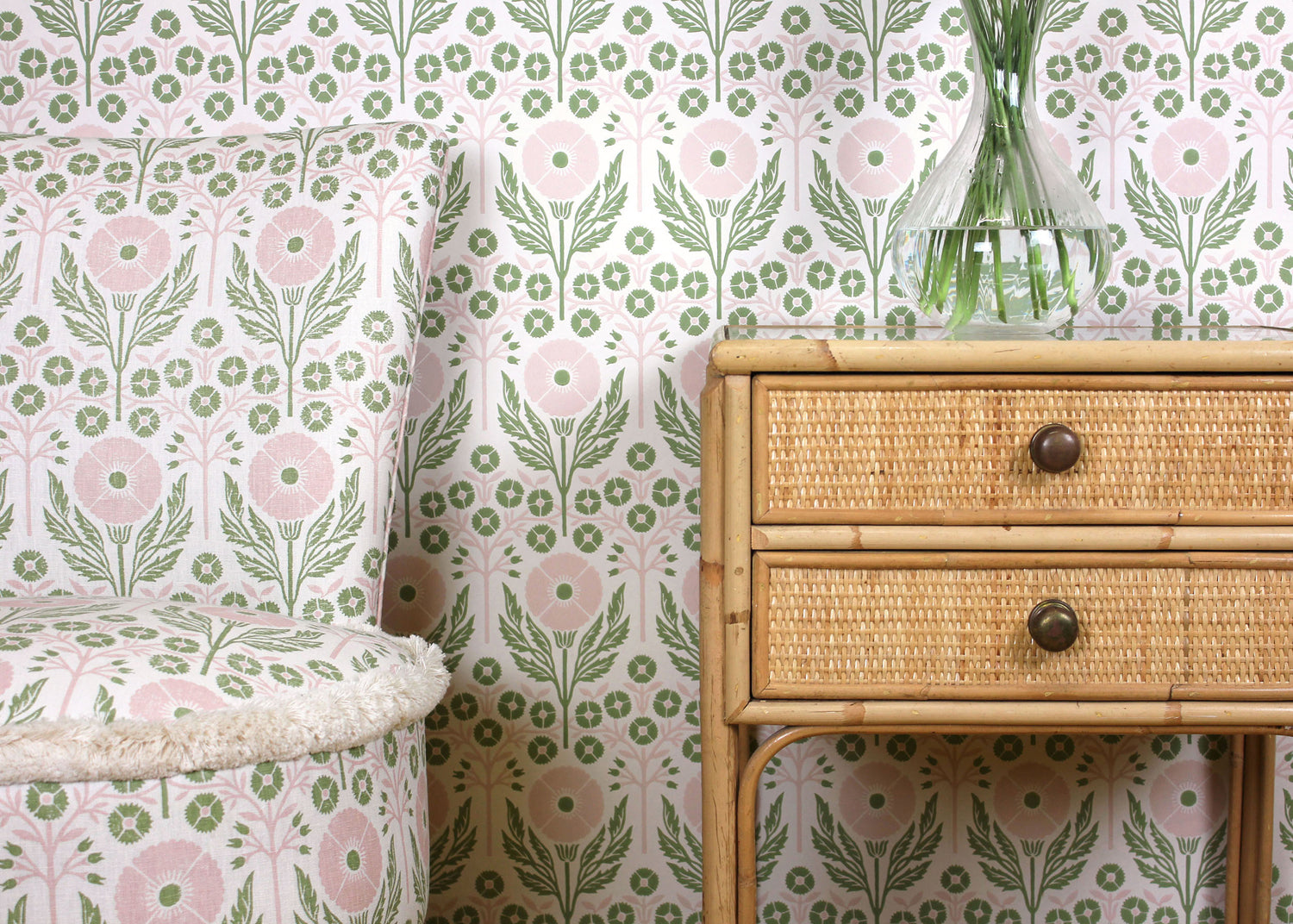 Upholstered side chair and wallpaper in matching pink and green floral pattern along side a rattan table with drawers.
