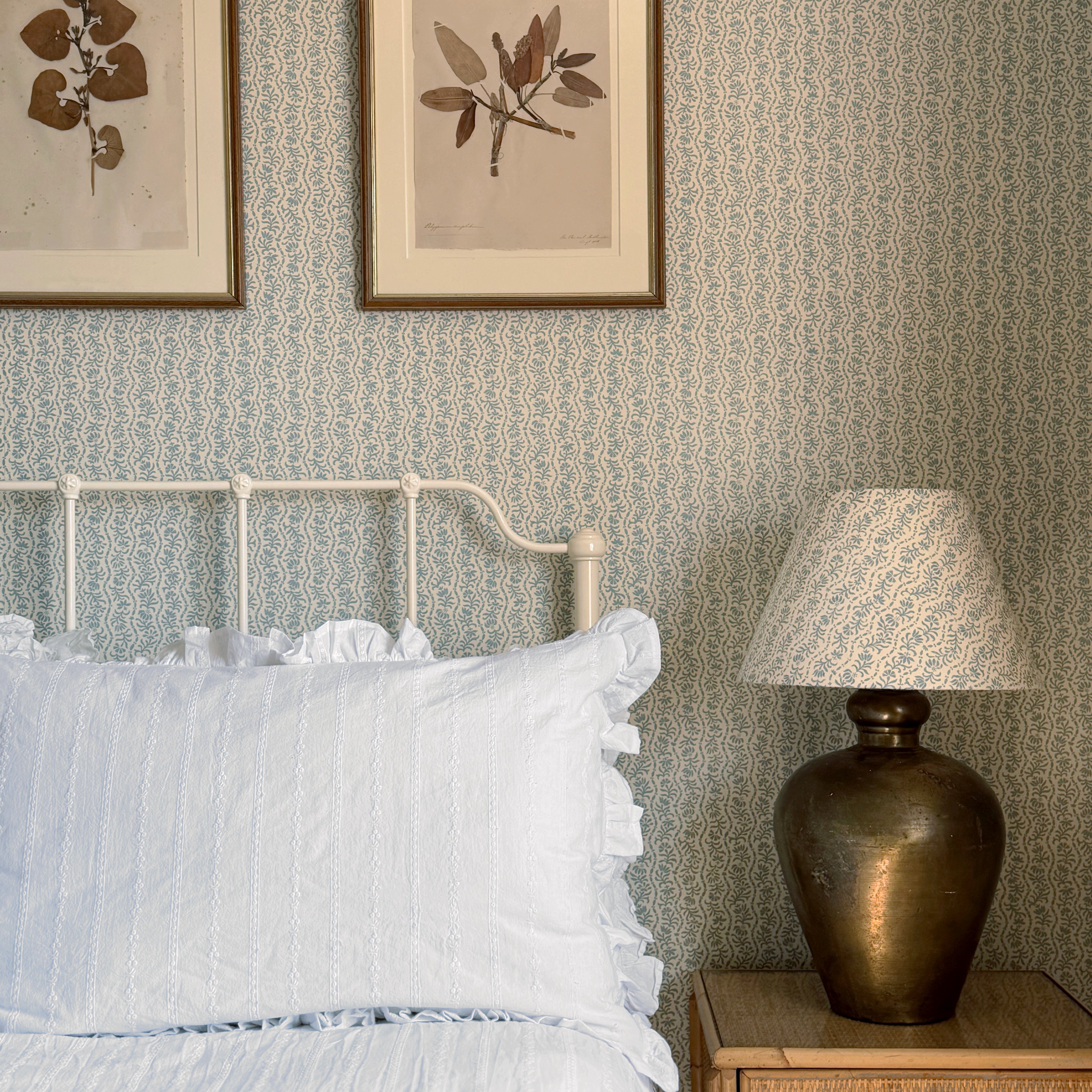 A styled bedroom corner featuring a white metal bed frame with white bedding and a mustard yellow throw, set against a light blue patterned wallpaper. A wooden nightstand with a bronze lamp sits on the right, and two framed botanical prints hang on the wall above the headboard.