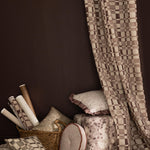 Detail of a styled interior scene with a floor to ceiling draped fabric with a brown and white checkerboard along side stacked throw pillows, ottoman and basket with wallpaper rolls in various patterns in a range of muted colors against a dark brown wall. 