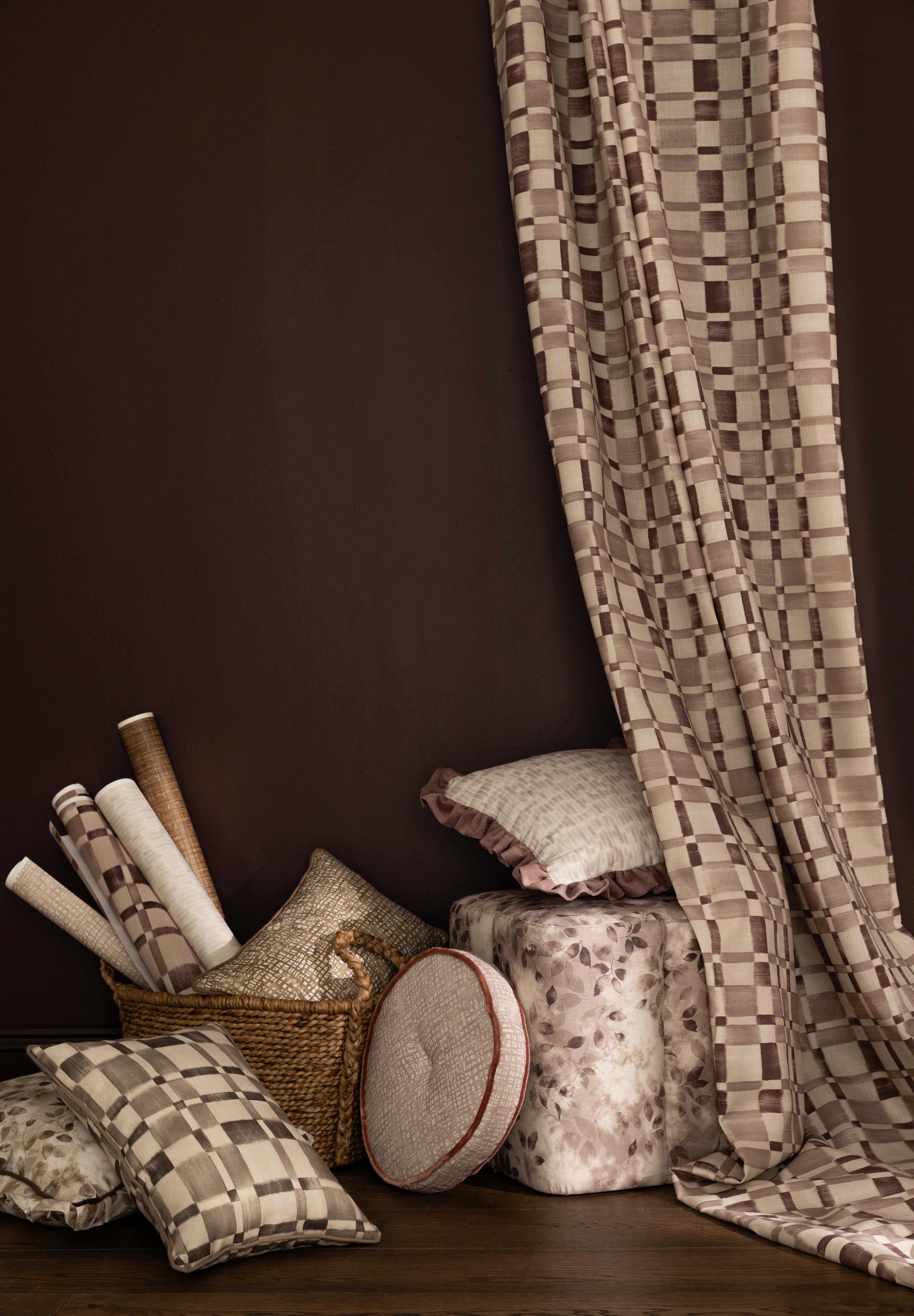 Detail of a styled interior scene with a floor to ceiling draped fabric with a brown and white checkerboard along side stacked throw pillows, ottoman and basket with wallpaper rolls in various patterns in a range of muted colors against a dark brown wall. 