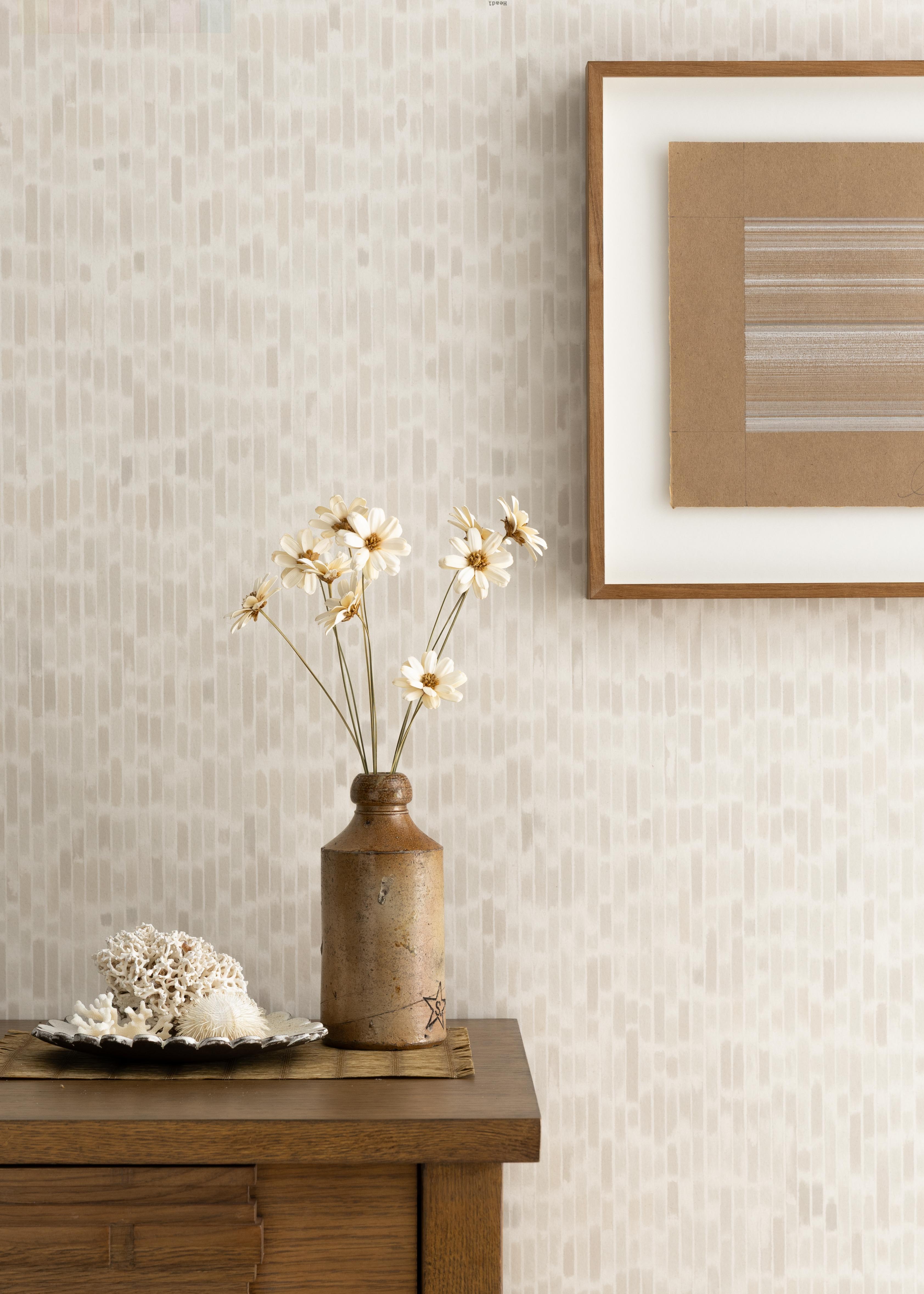 Detail of a styled interior scene set against wallpaper with a soft beige a watercolor broken stripe pattern featuring a wooden dresser, ceramic vase with daisies, dish with seashells against a wall with a framed brown and white abstract line drawing. 