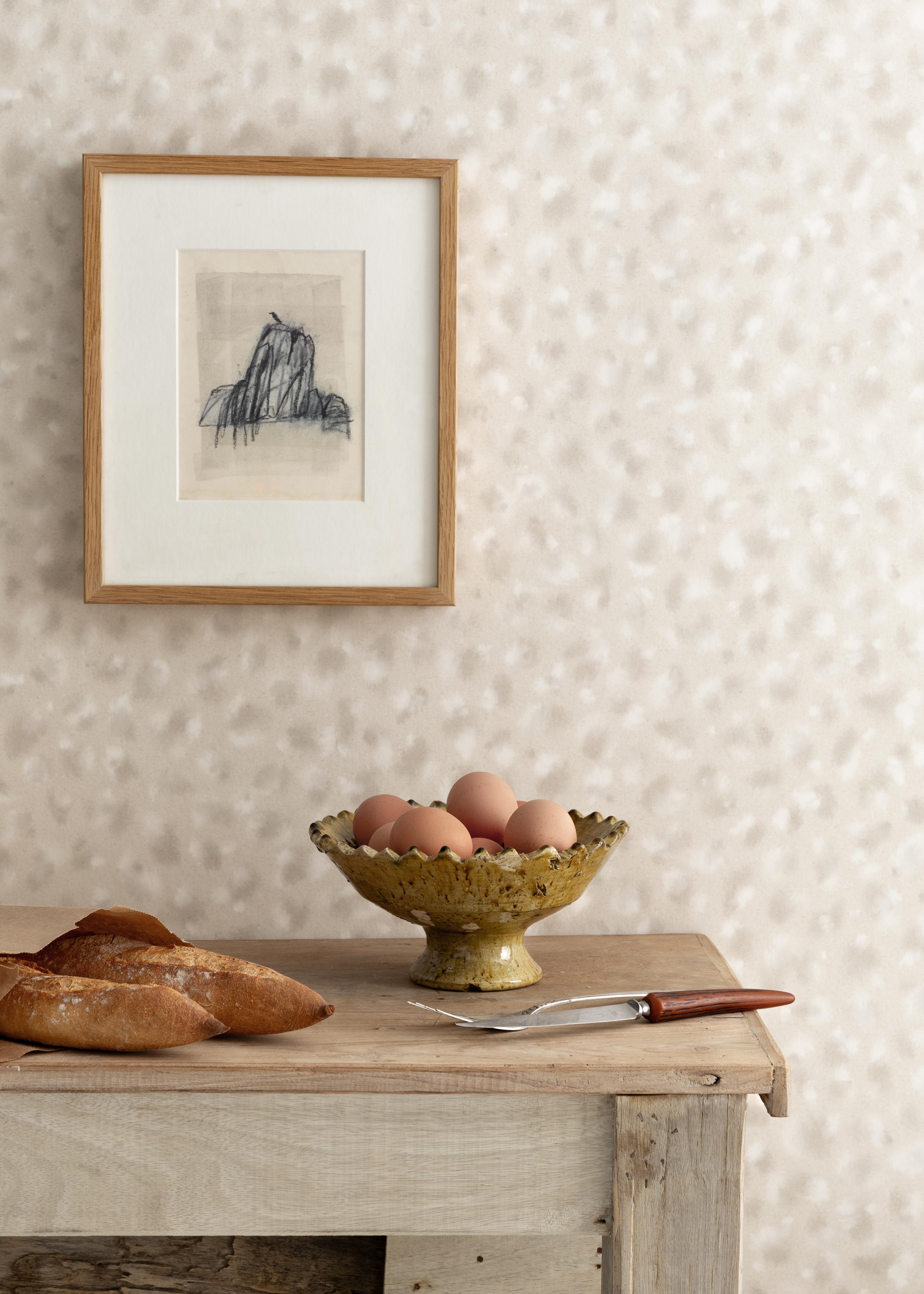 Detail of a styled interior scene set against wallpaper in beige watercolor animal patter with a framed abstract charcoal sketch with a rustic wooden table with two baguettes, footed ceramic bowl with brown eggs, serving fork and knife.