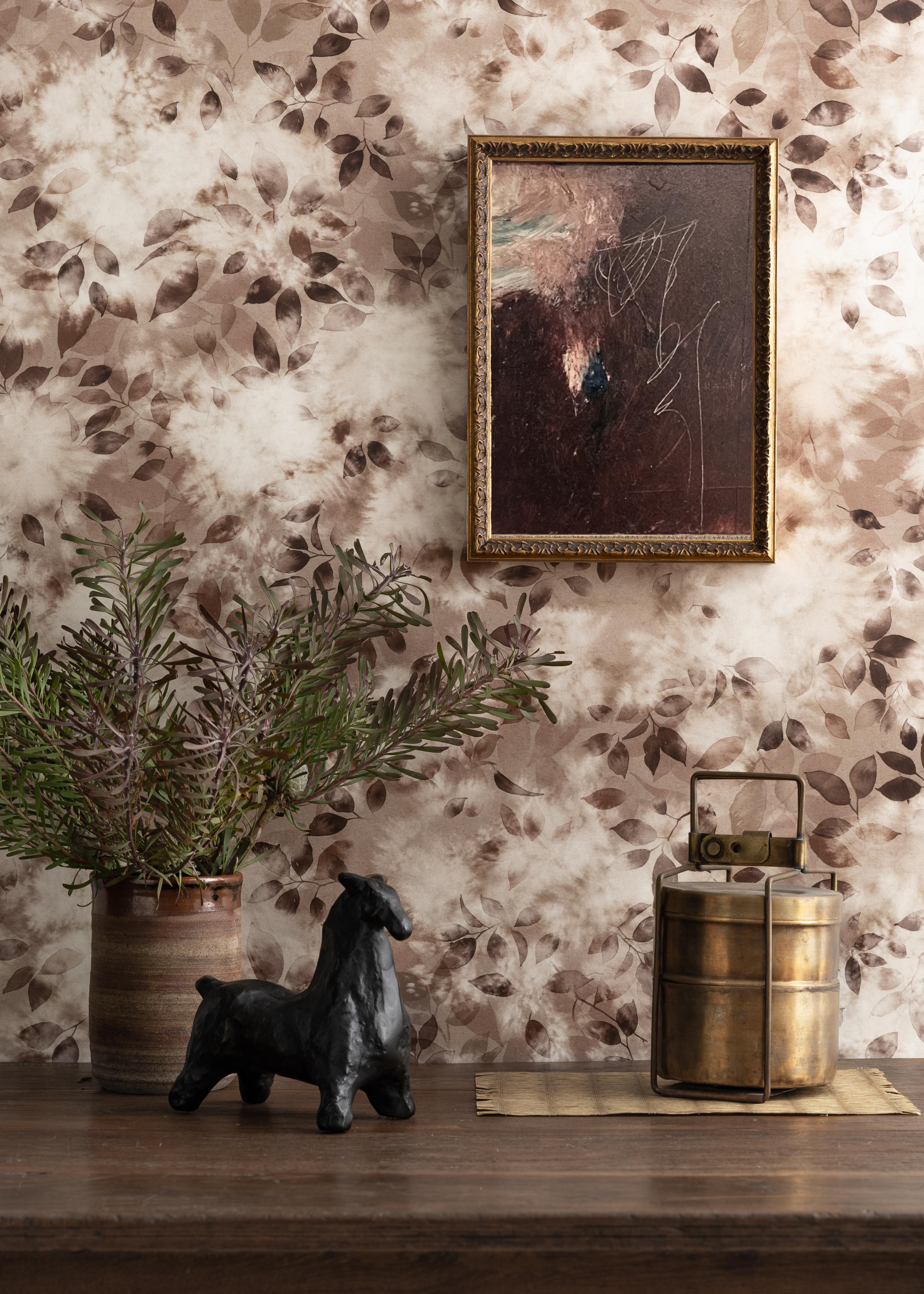 Detail of a styled interior scene set against wallpaper in a brown watercolor leaf pattern with a framed abstract painting featuring a wooden table with a ceramic vase with greenery and a black ceramic horse sculpture and a brass canteen and rattan placemat. 