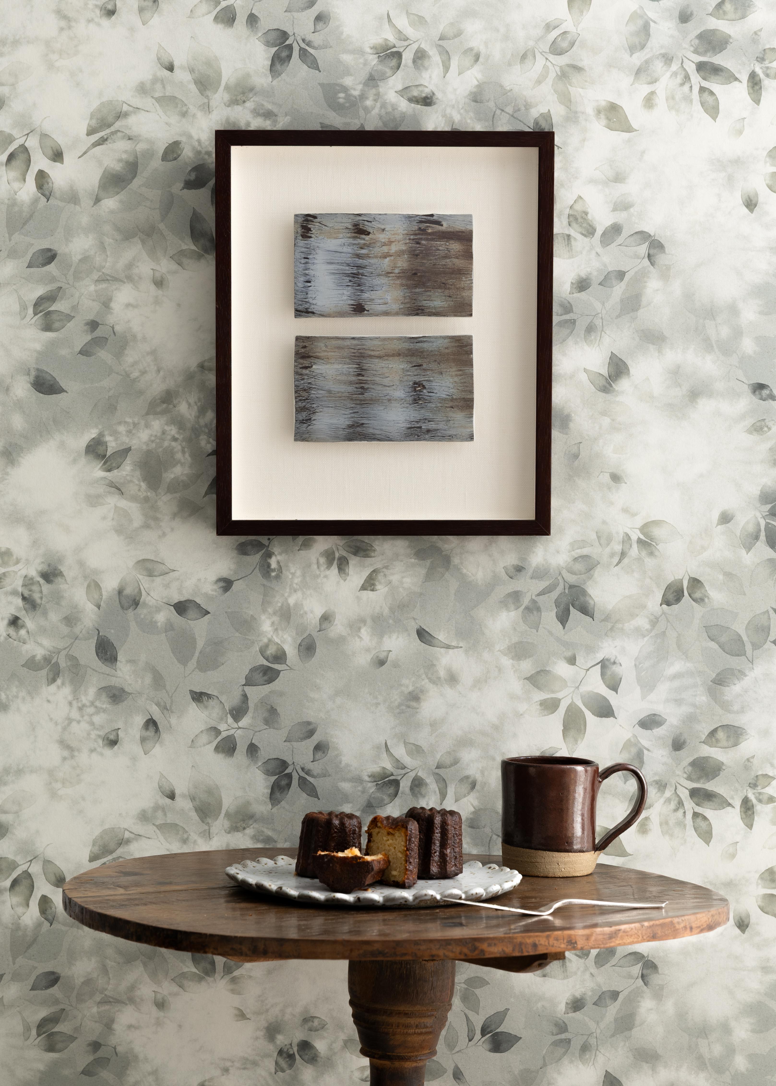 Detail of a styled interior scene set against wallpaper with a grey watercolor leaf pattern featuring a framed stacked abstract prints and an antique wooden table with ceramic mug and plate with pastries and a serving spoon. 
