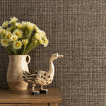 Detail of a styled interior scene set against wallpaper in a grey textured check with wooden dresser, ceramic picture with yellow flowers and an antique painted wooden carving of a goose. 