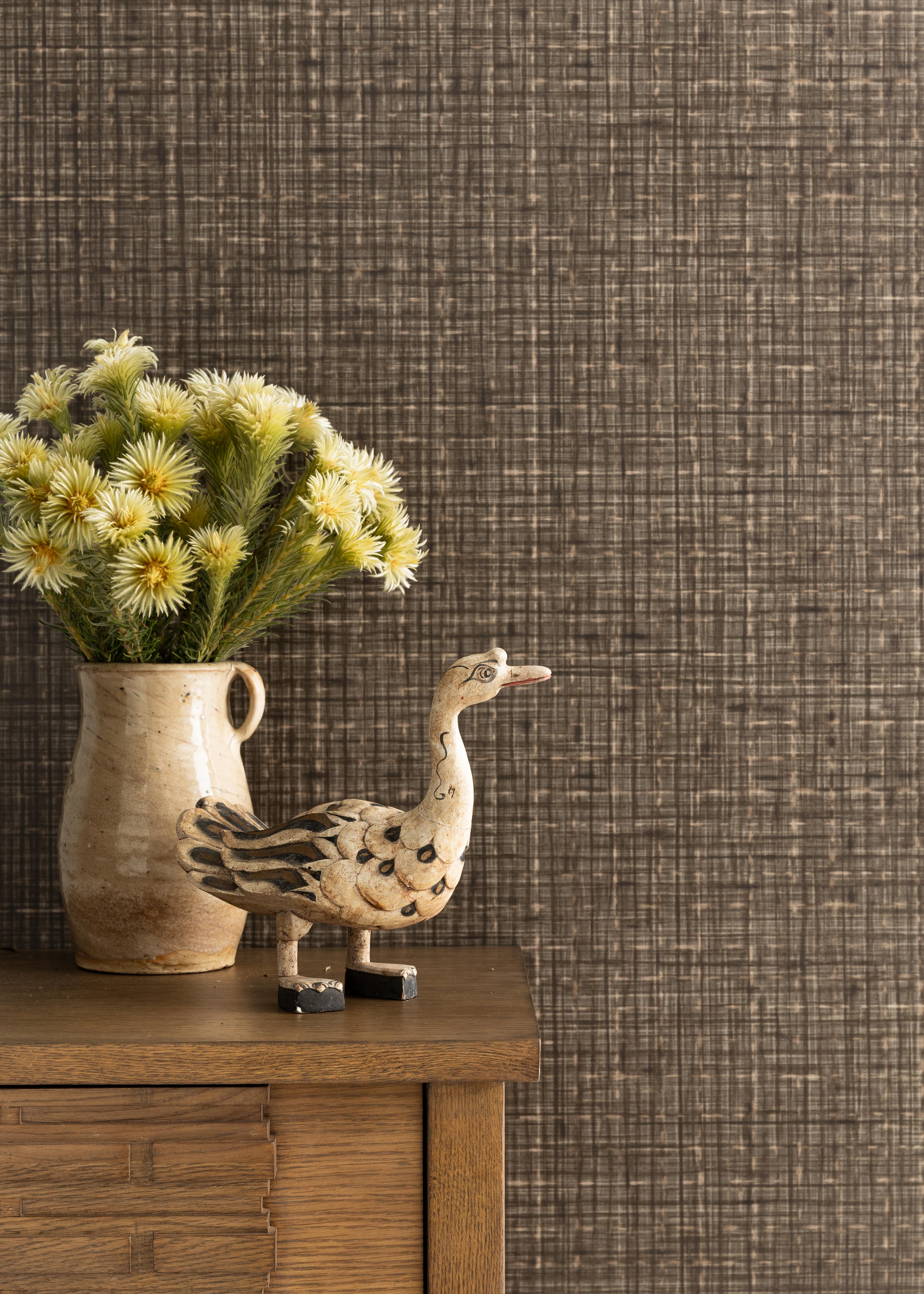 Detail of a styled interior scene set against wallpaper in a grey textured check with wooden dresser, ceramic picture with yellow flowers and an antique painted wooden carving of a goose. 