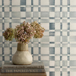 Detail of a styled interior scene set against wallpaper in a blue and white checkerboard pattern with a wooden dresser with dried flowers in a ceramic vase stacked on hardcover books. 