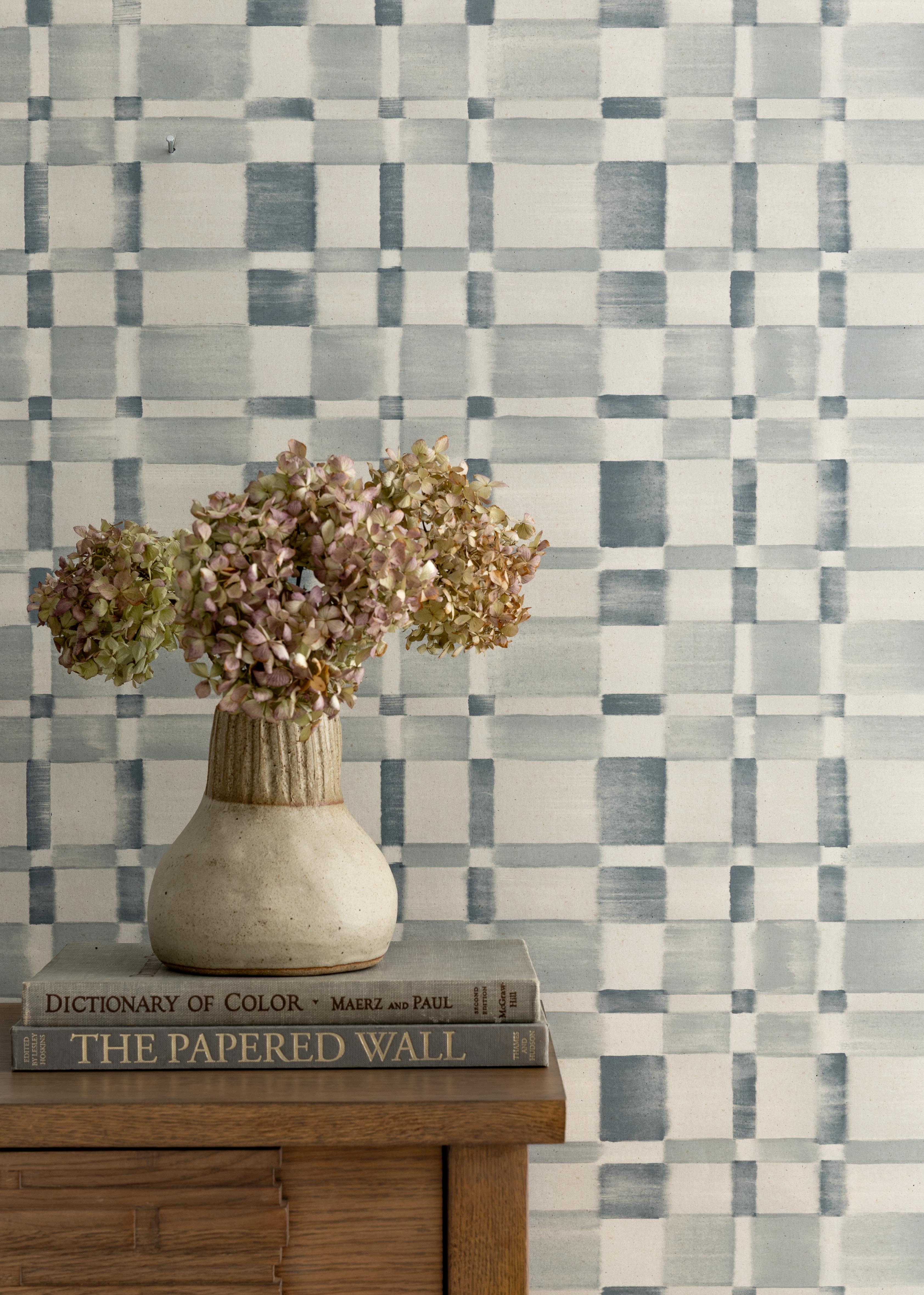 Detail of a styled interior scene set against wallpaper in a blue and white checkerboard pattern with a wooden dresser with dried flowers in a ceramic vase stacked on hardcover books. 