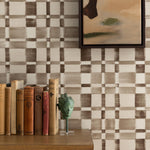 Detail of a styled interior scene set against wallpaper in a charcoal grey checkerboard design with a framed abstract panting and a wooden dresser with antique hardcover books and bookend with a metal sculpture of a woman's head. 