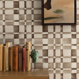 Detail of a styled interior scene set against wallpaper in a charcoal grey checkerboard design with a framed abstract panting and a wooden dresser with antique hardcover books and bookend with a metal sculpture of a woman's head. 
