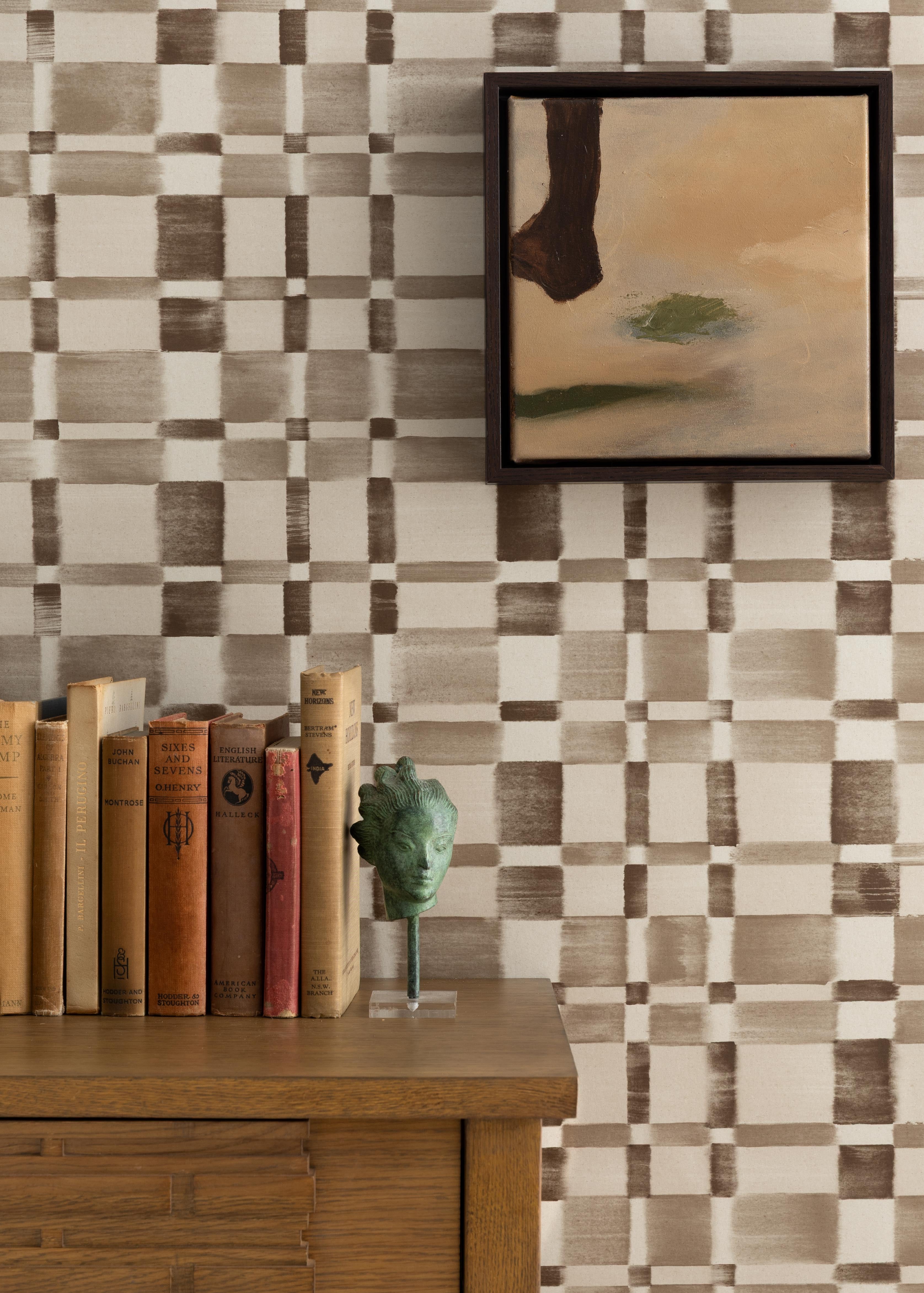 Detail of a styled interior scene set against wallpaper in a charcoal grey checkerboard design with a framed abstract panting and a wooden dresser with antique hardcover books and bookend with a metal sculpture of a woman's head. 