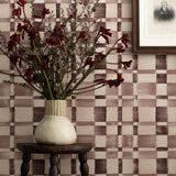 Detail of a styled interior scene set against wallpaper with a wine purple checkerboard design featuring a framed antique portrait of a soldier and a dark wood stool with carved legs and a ceramic vase with red flowers and dried branches.