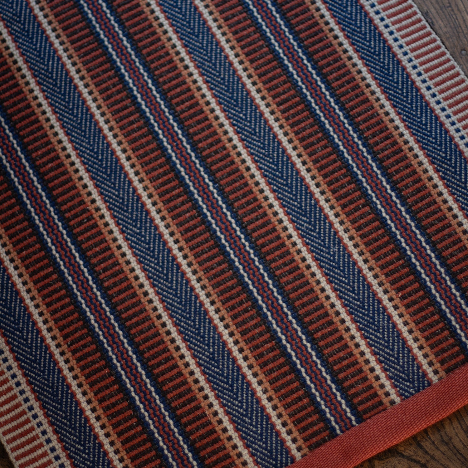 Detail of an area rug on a wood floor in a complex woven stripe made of wool in shades of blue and red. 