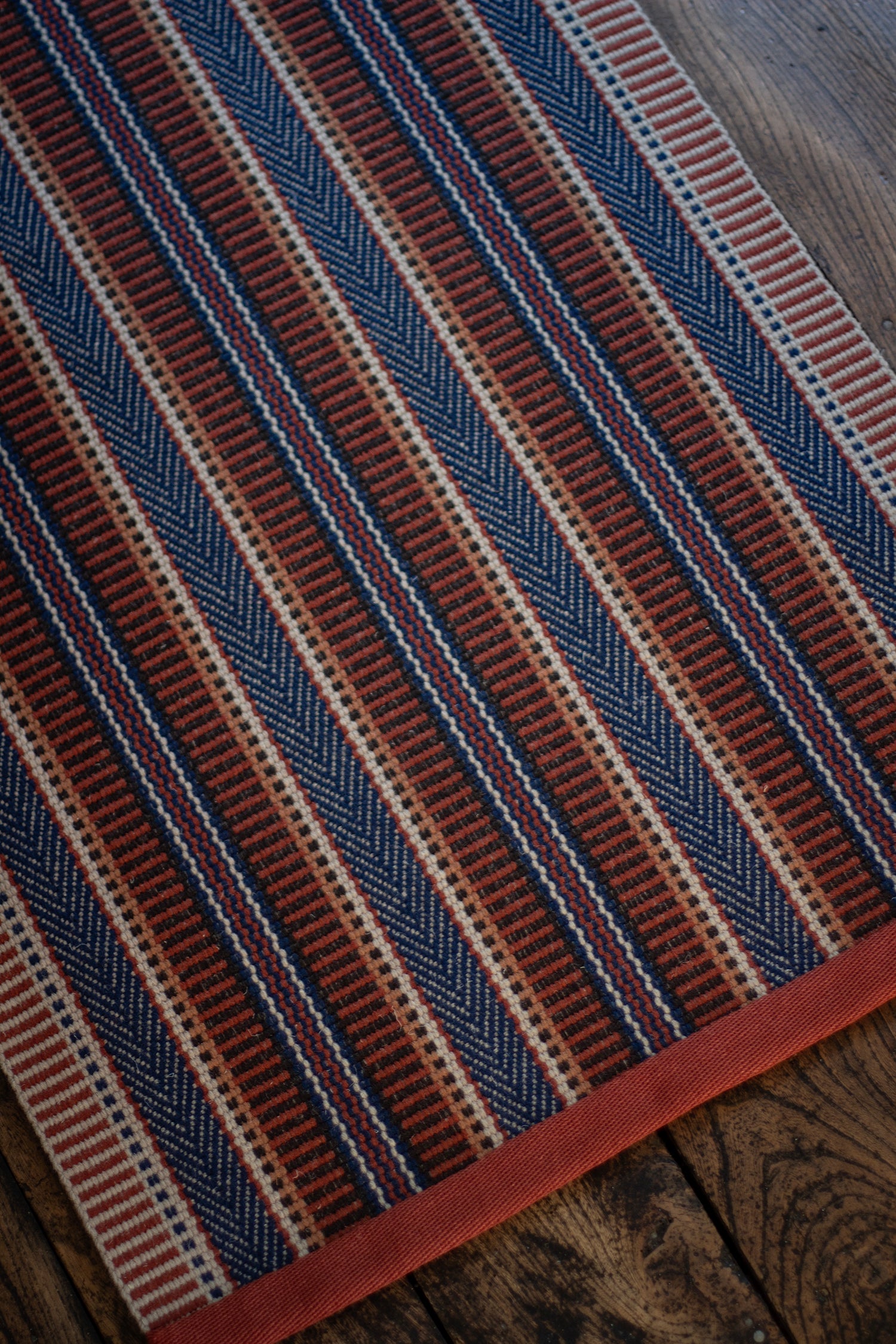 Detail of an area rug on a wood floor in a complex woven stripe made of wool in shades of blue and red. 