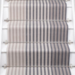 A close-up view of a staircase runner with vertical gray and beige herringbone stripes, mounted on white stairs with metal rails.