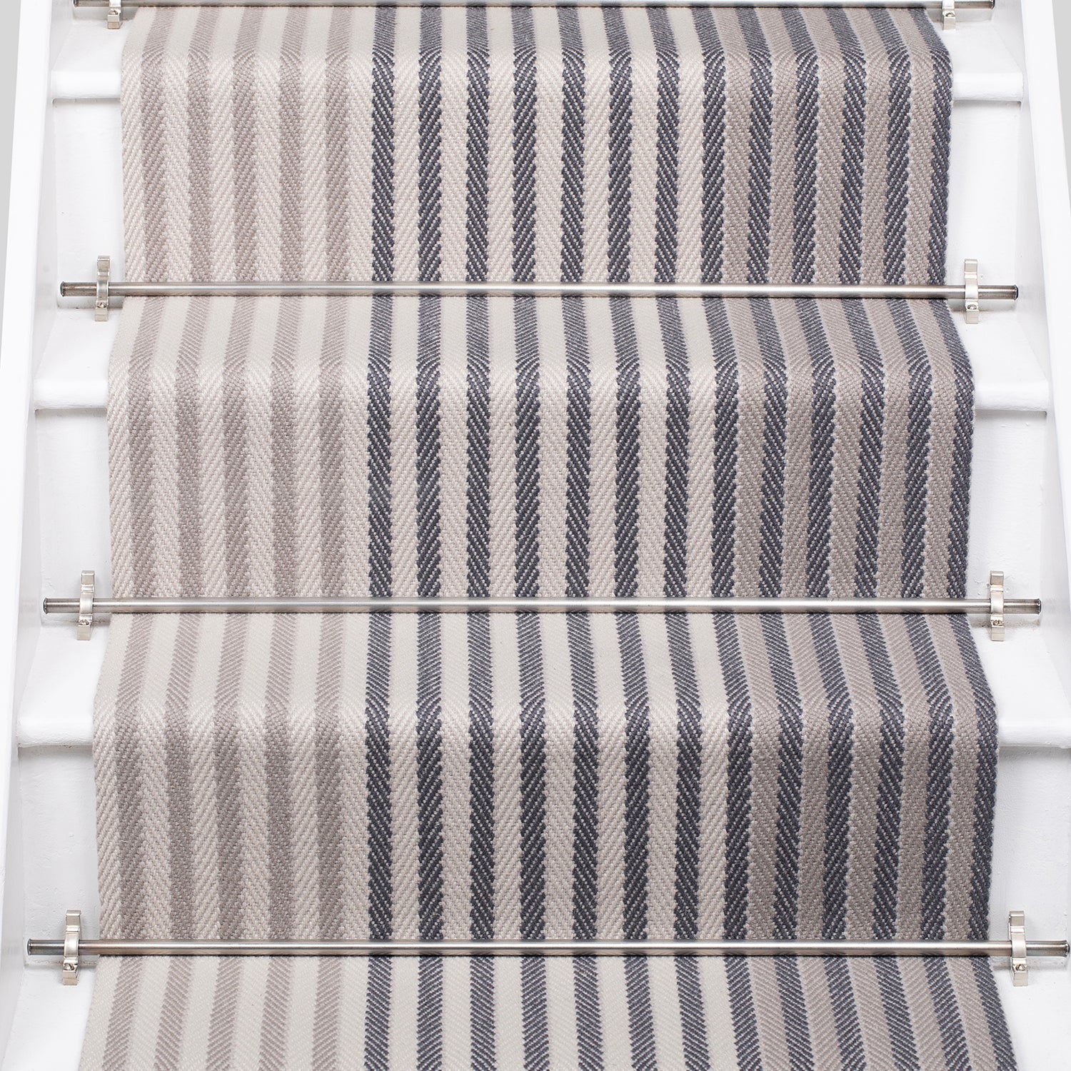 A close-up view of a staircase runner with vertical gray and beige herringbone stripes, mounted on white stairs with metal rails.