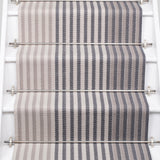 A close-up view of a staircase runner with vertical gray and beige herringbone stripes, mounted on white stairs with metal rails.