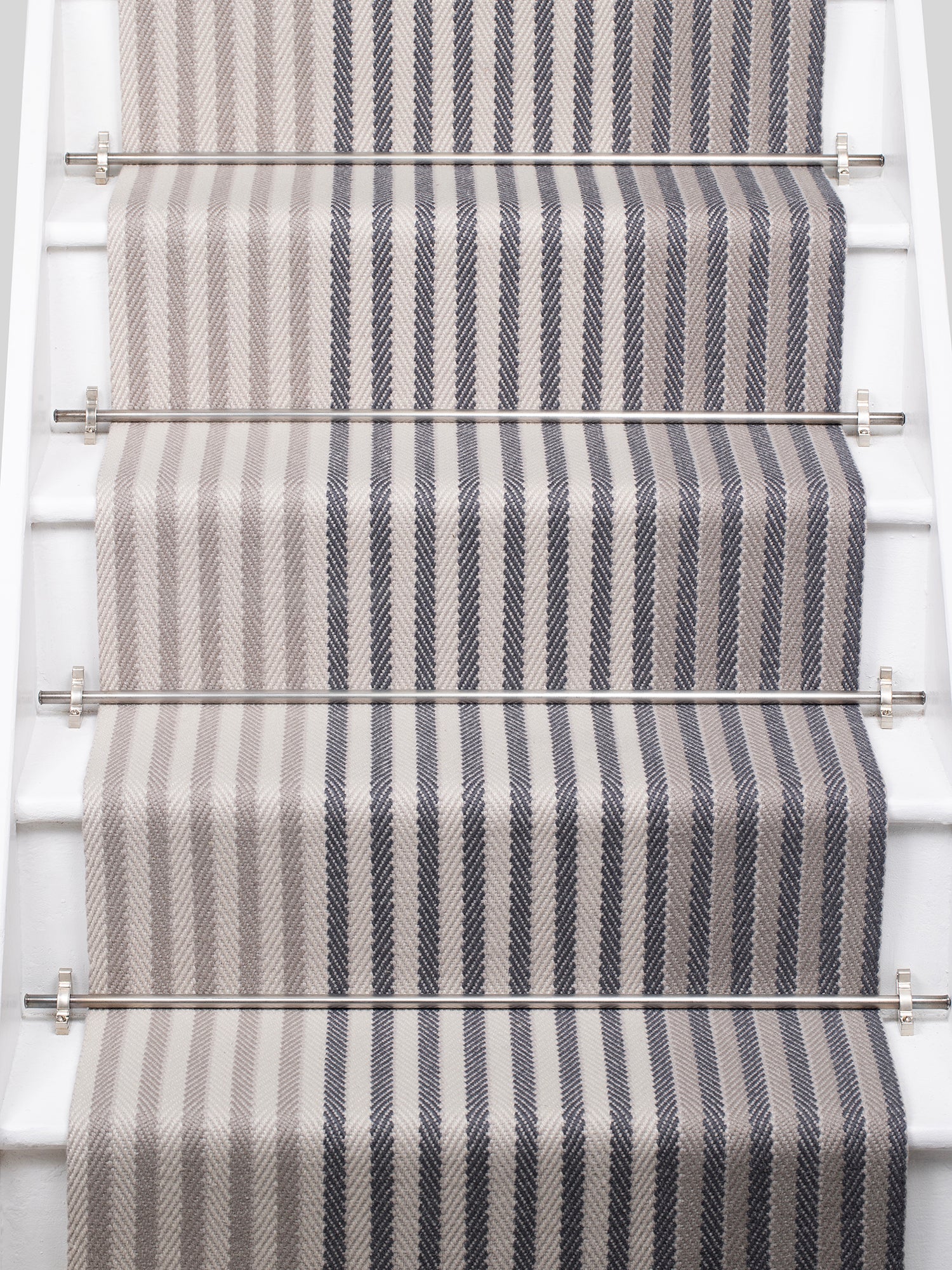 A close-up view of a staircase runner with vertical gray and beige herringbone stripes, mounted on white stairs with metal rails.