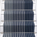 A close-up view of a staircase runner with vertical navy blue, steel and gray herringbone stripes, mounted on white stairs with metal rails.
