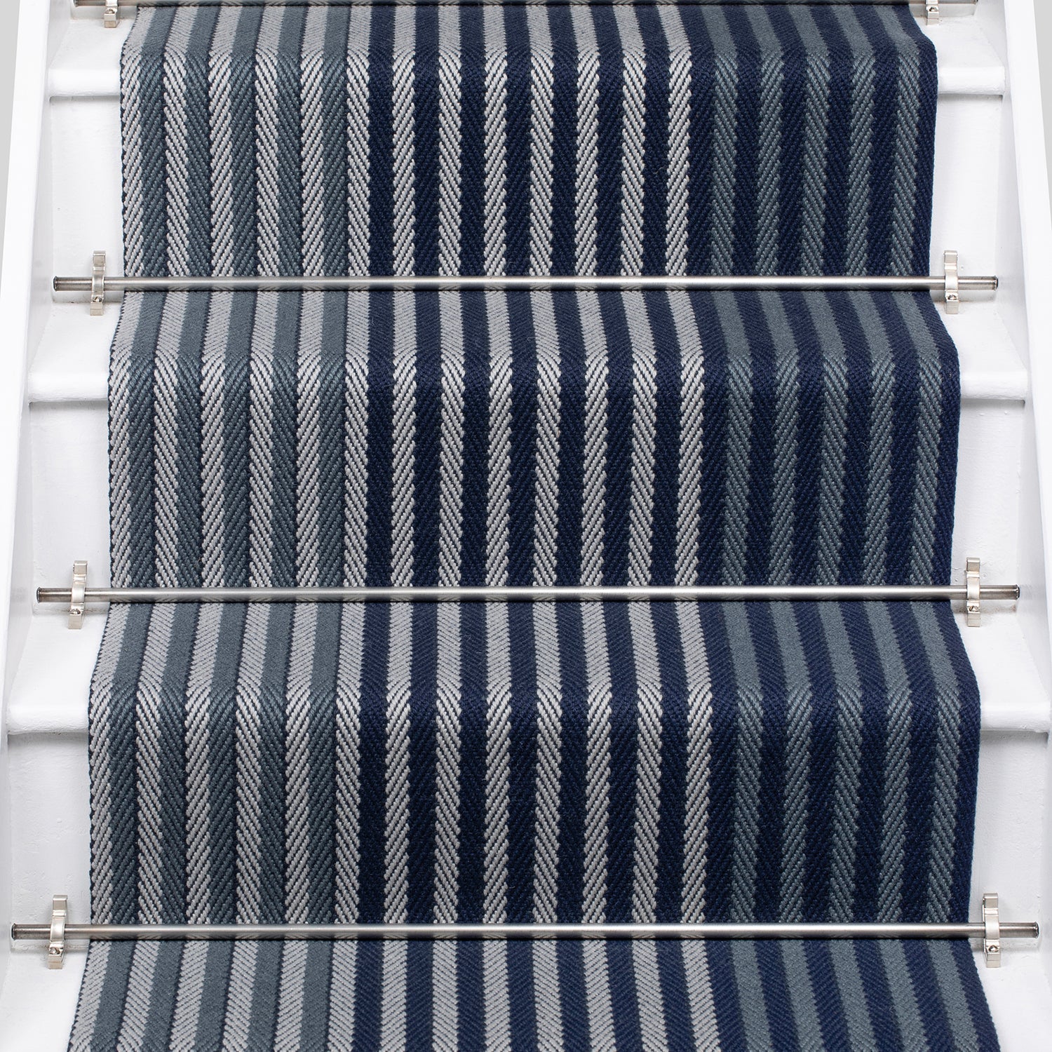 A close-up view of a staircase runner with vertical navy blue, steel and gray herringbone stripes, mounted on white stairs with metal rails.
