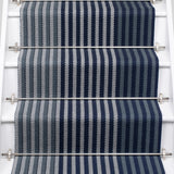 A close-up view of a staircase runner with vertical navy blue, steel and gray herringbone stripes, mounted on white stairs with metal rails.
