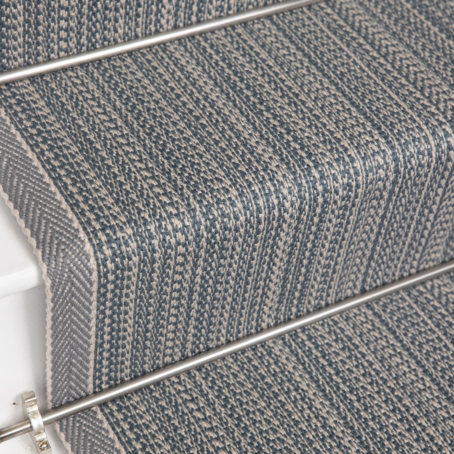 Close-up of carpeted stairs with a textured herringbone pattern in gray and blue tones, showing the stair treads and metal rail.