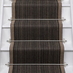 A close-up view of a stair runner carpet installed on a staircase, featuring a textured herringbone pattern in black, brown, and beige with silver metal rails.