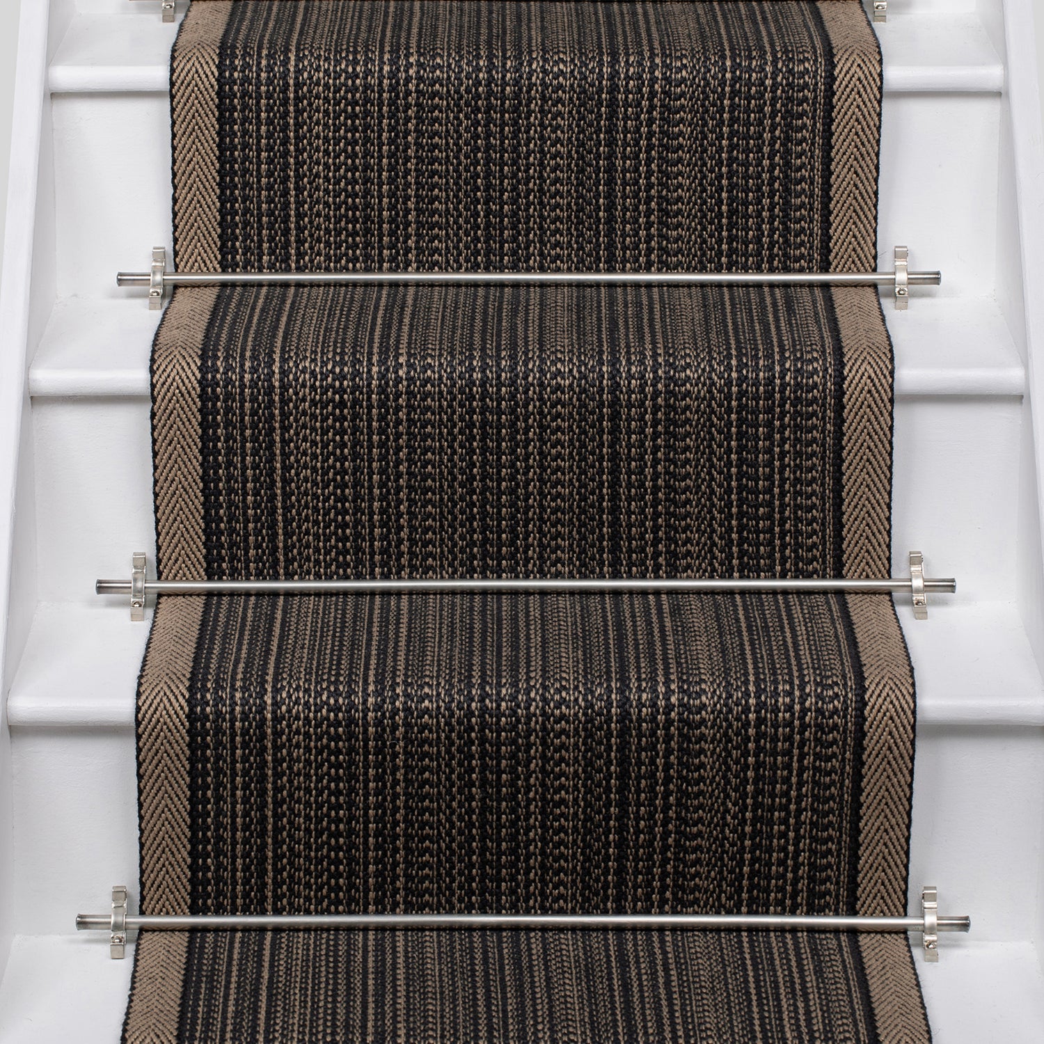 A close-up view of a stair runner carpet installed on a staircase, featuring a textured herringbone pattern in black, brown, and beige with silver metal rails.