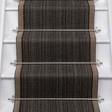 A close-up view of a stair runner carpet installed on a staircase, featuring a textured herringbone pattern in black, brown, and beige with silver metal rails.
