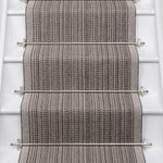 A close-up view of a woven stair runner installed on a staircase, featuring a textured herringbone pattern in gray and beige with stainless steel rails.