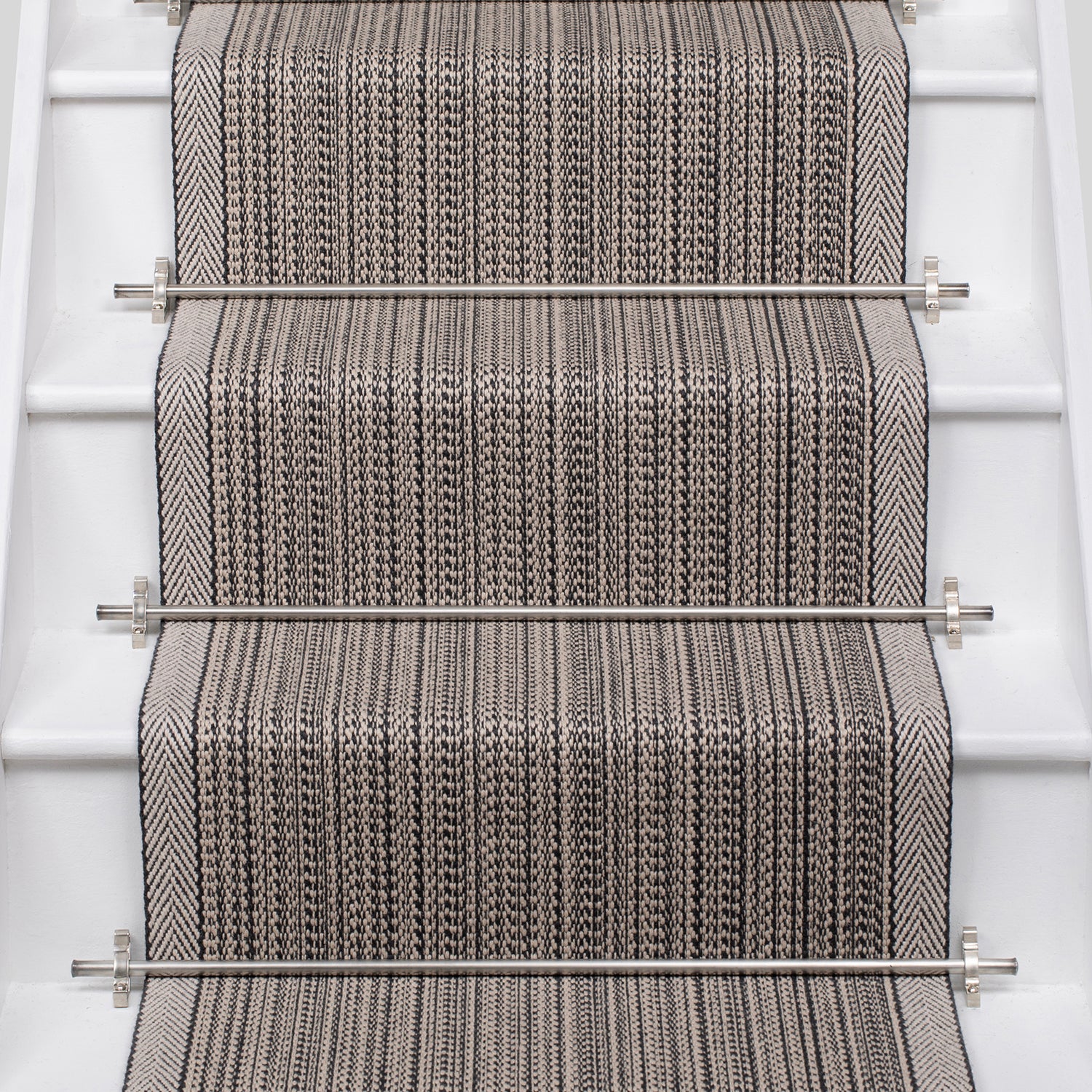 A close-up view of a woven stair runner installed on a staircase, featuring a textured herringbone pattern in gray and beige with stainless steel rails.
