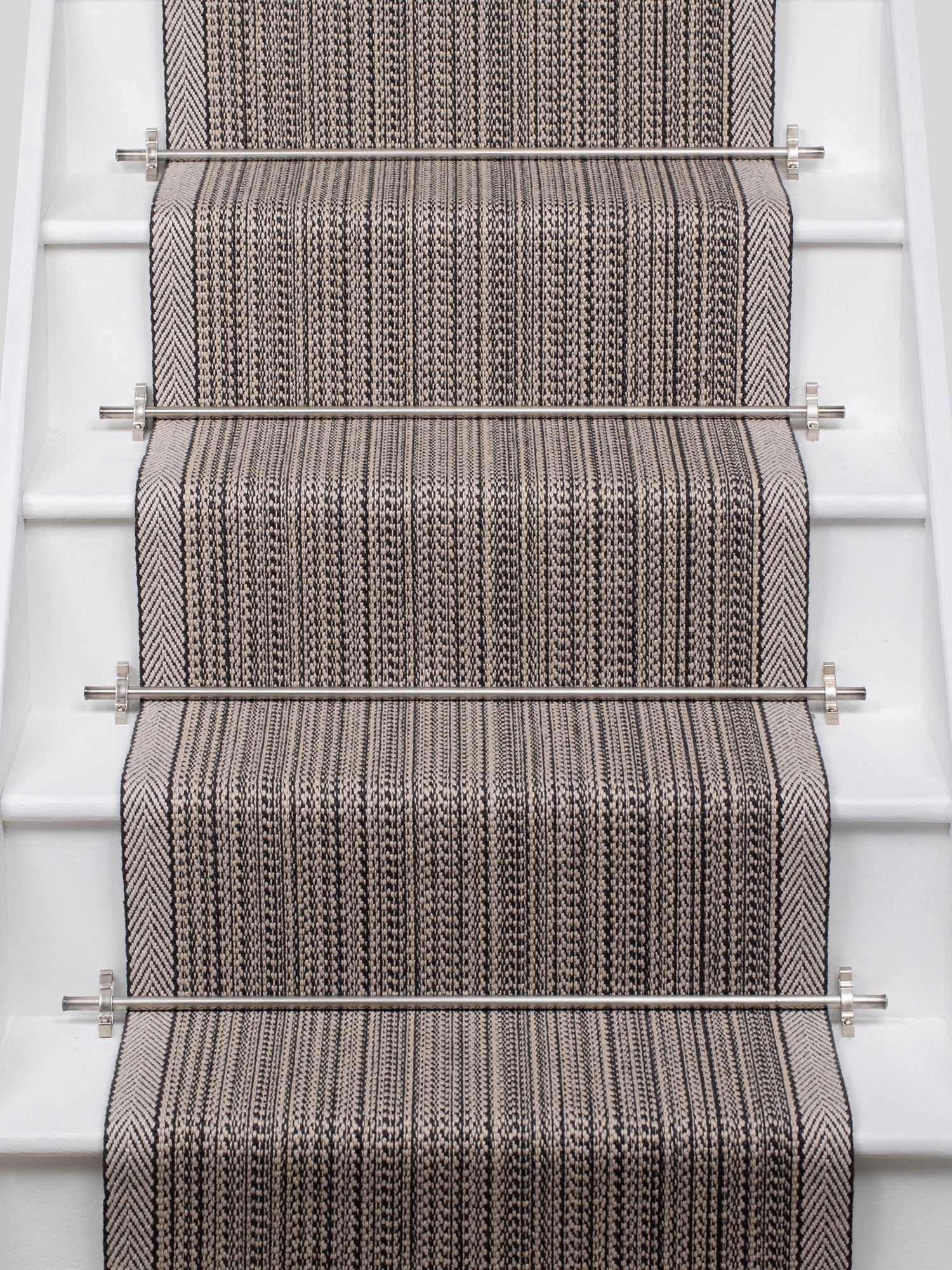 A close-up view of a woven stair runner installed on a staircase, featuring a textured herringbone pattern in gray and beige with stainless steel rails.
