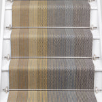 A close-up view of a staircase runner with vertical degradé stripes ranging from beige to yellow to navy blue mounted on white stairs with metal rails.