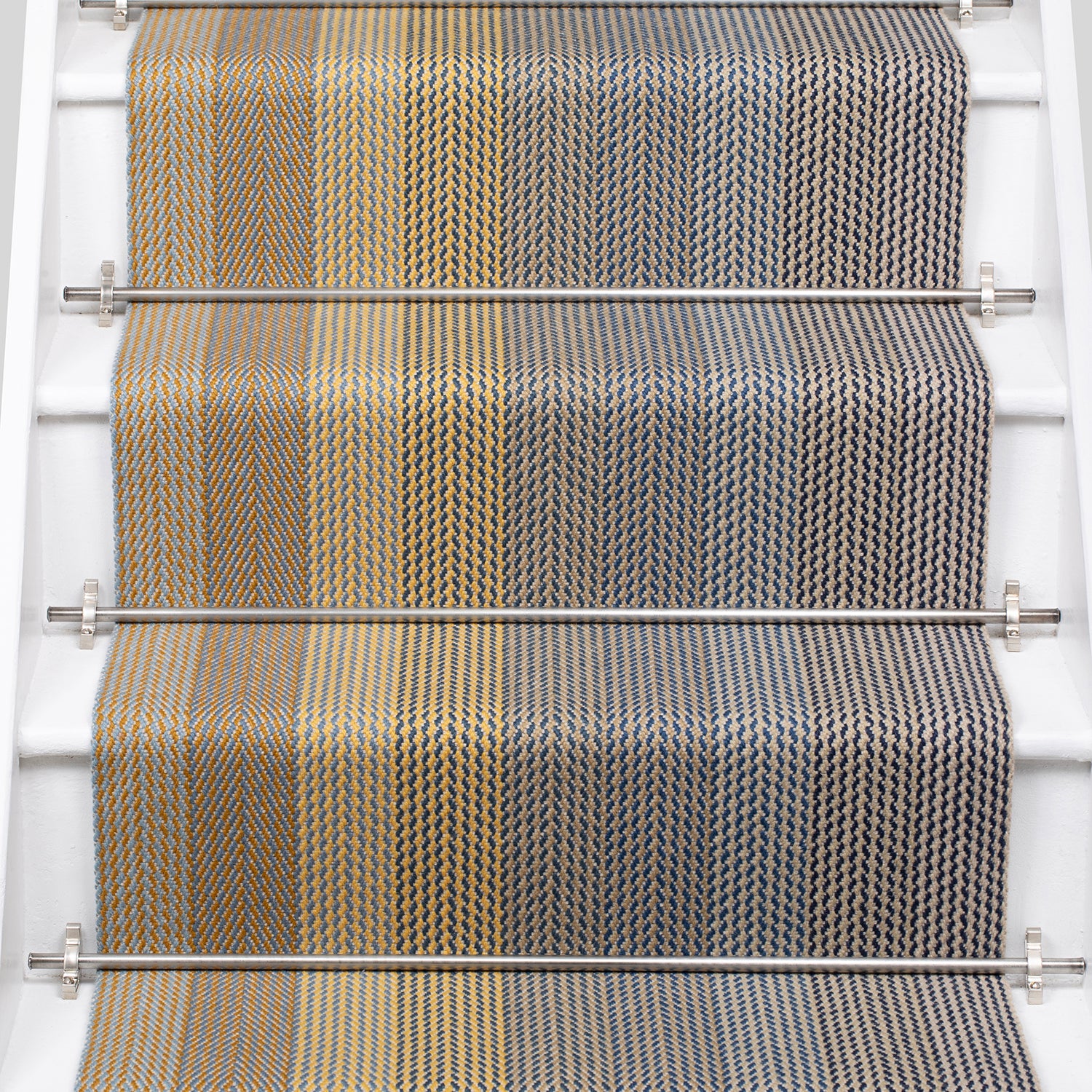 A close-up view of a staircase runner with vertical degradé stripes ranging from beige to yellow to navy blue mounted on white stairs with metal rails.