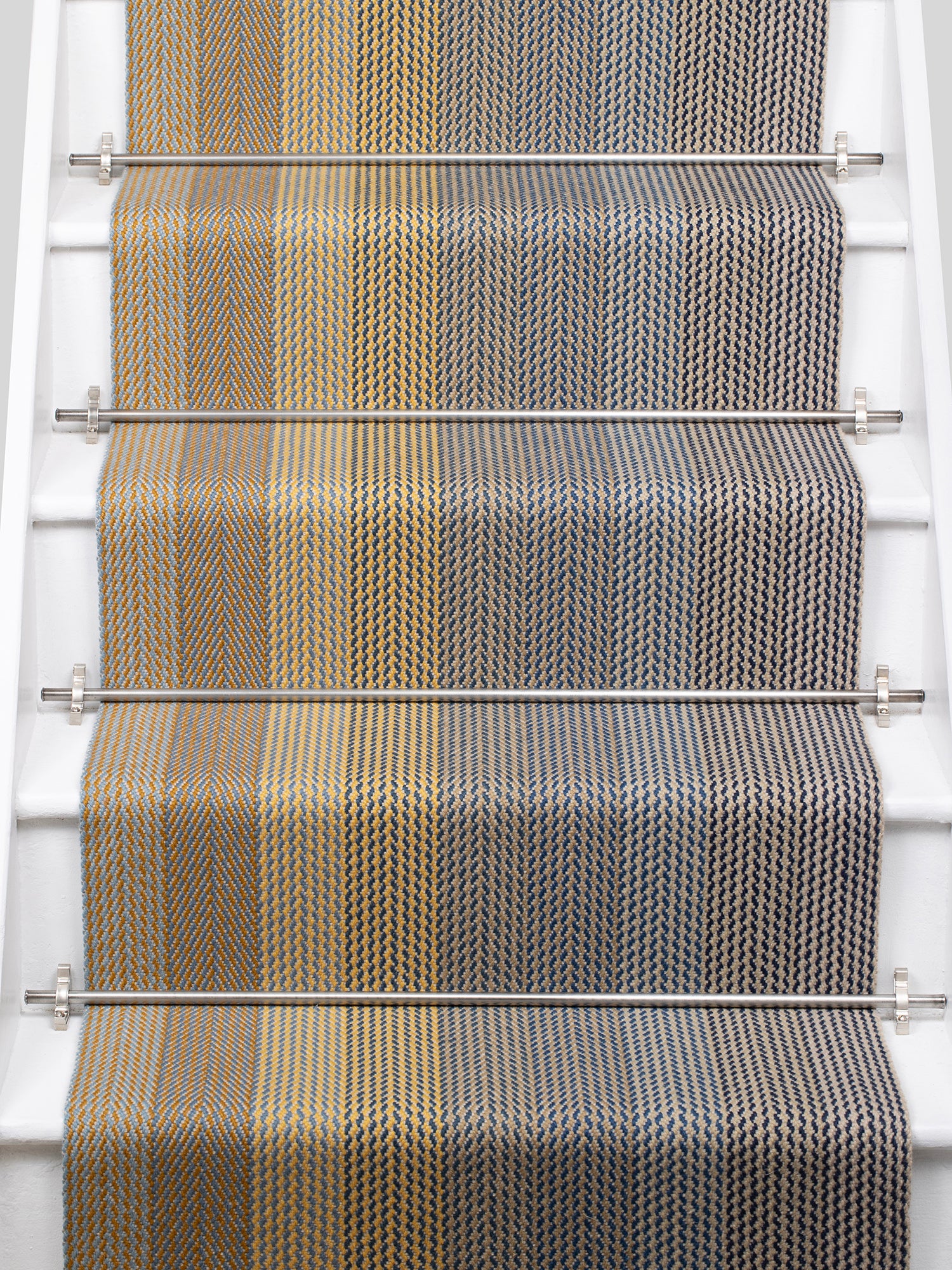 A close-up view of a staircase runner with vertical degradé stripes ranging from beige to yellow to navy blue mounted on white stairs with metal rails.