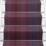 A close-up view of a staircase runner with vertical degradé stripes ranging from deep burgundy, purple, to blue mounted on white stairs with metal rails. 