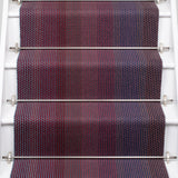A close-up view of a staircase runner with vertical degradé stripes ranging from deep burgundy, purple, to blue mounted on white stairs with metal rails. 