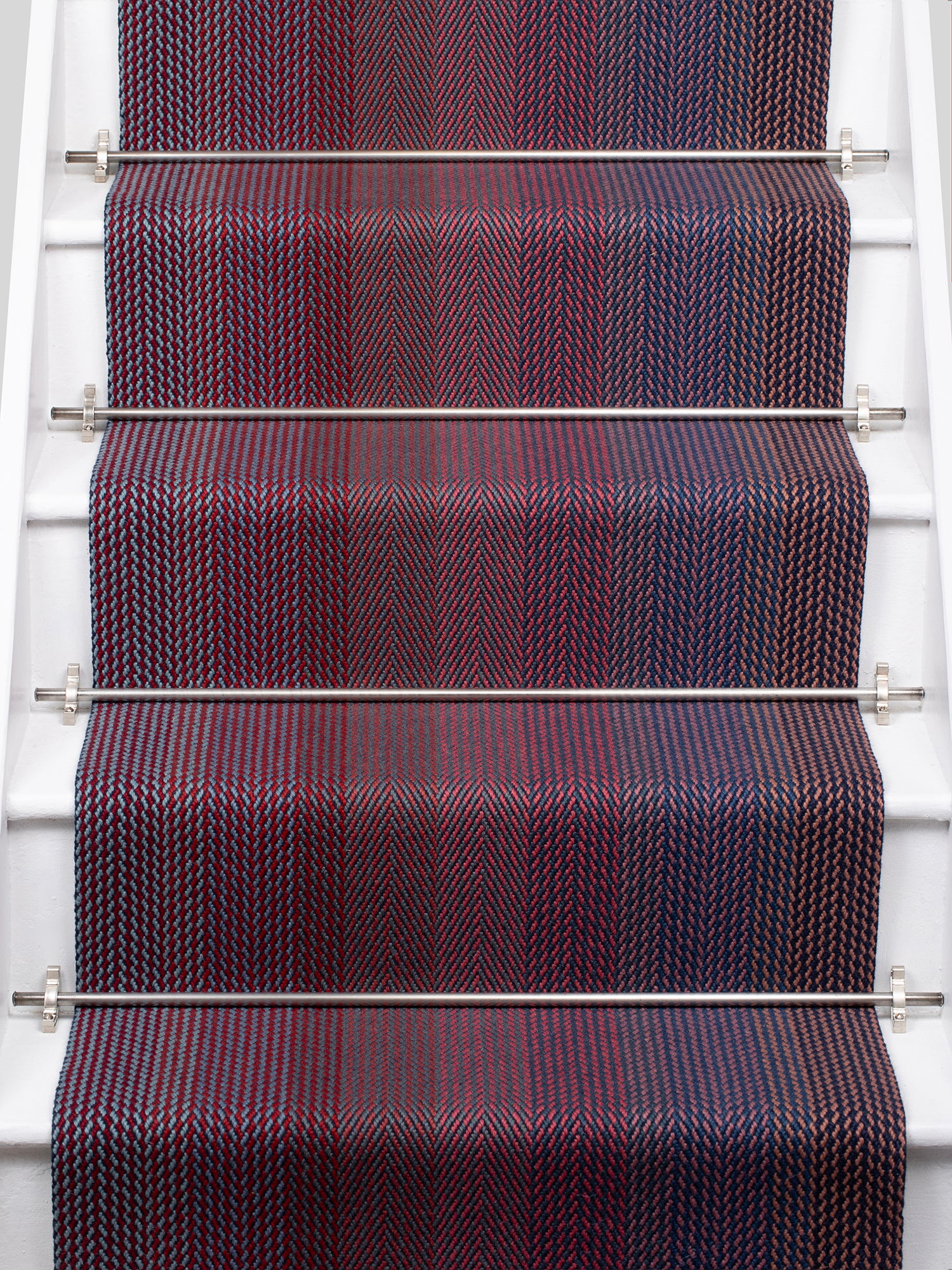 A close-up view of a staircase runner with vertical degradé stripes ranging from deep burgundy, purple, to blue mounted on white stairs with metal rails. 