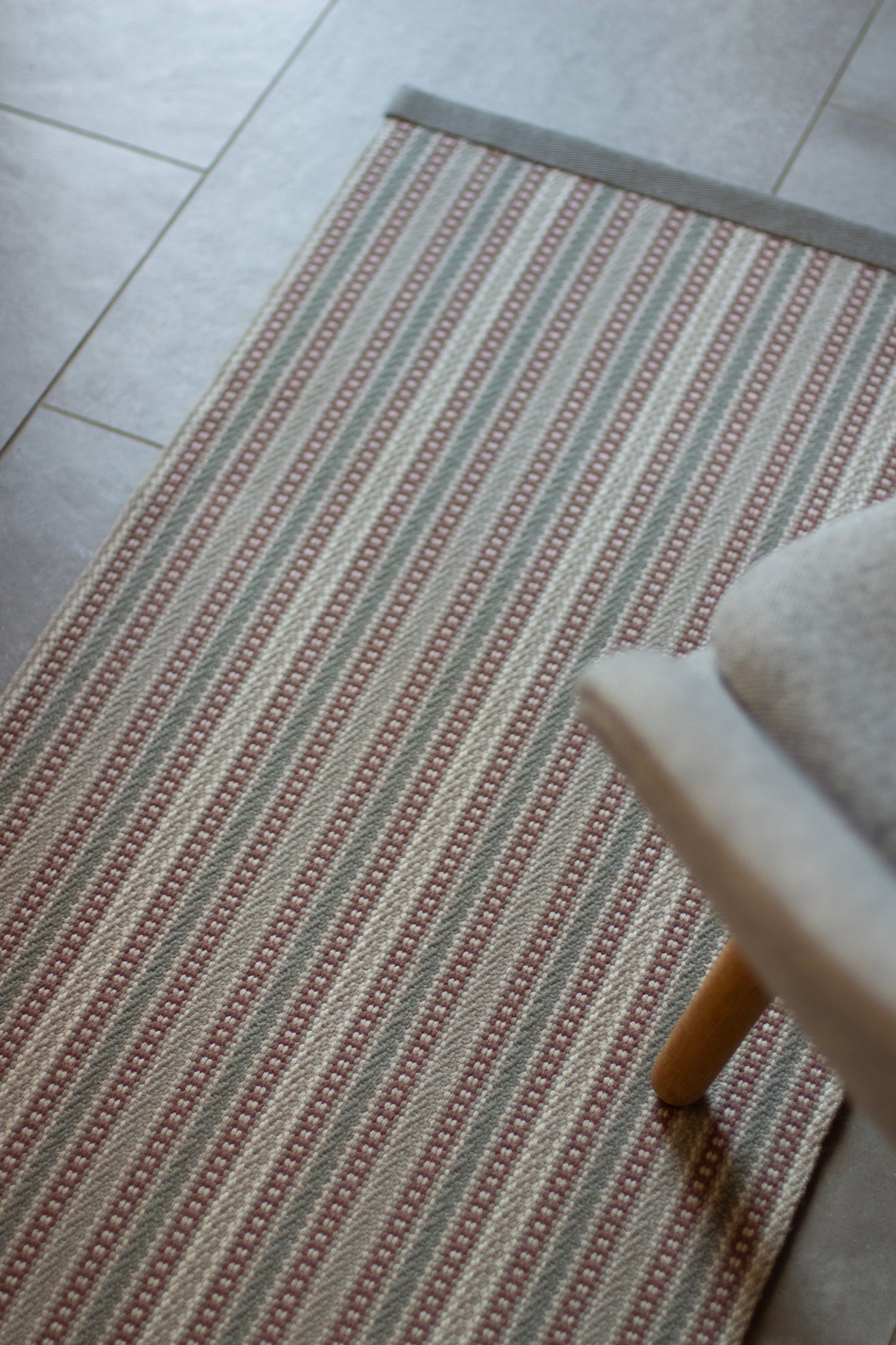 Detail of an area rug on a tile floor in a complex woven stripe design of wool in shades of pink and grey.