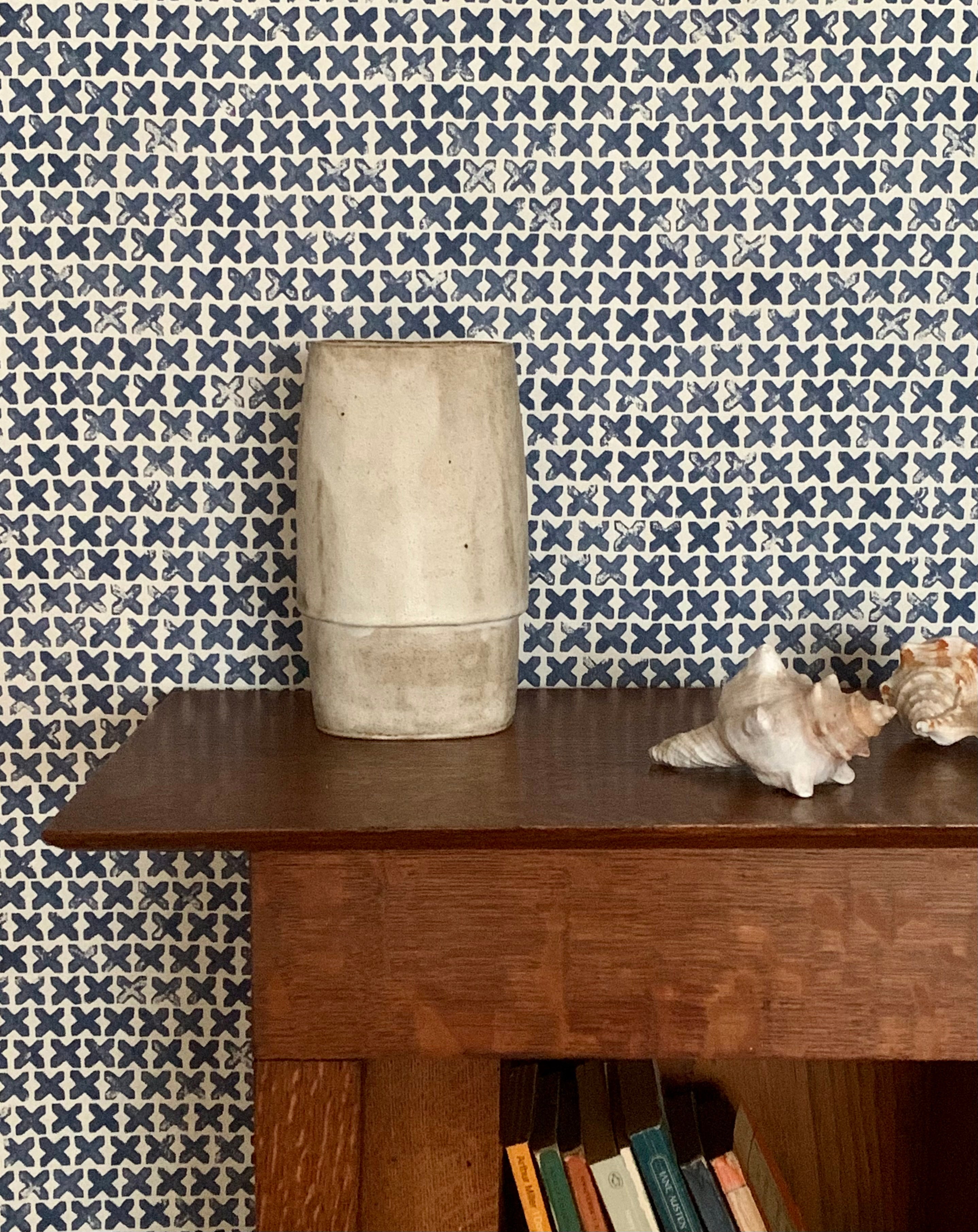 Wooden bookslef with a ceramic vase and conch shells against a blue and cream 'x' patterned wallpaper 