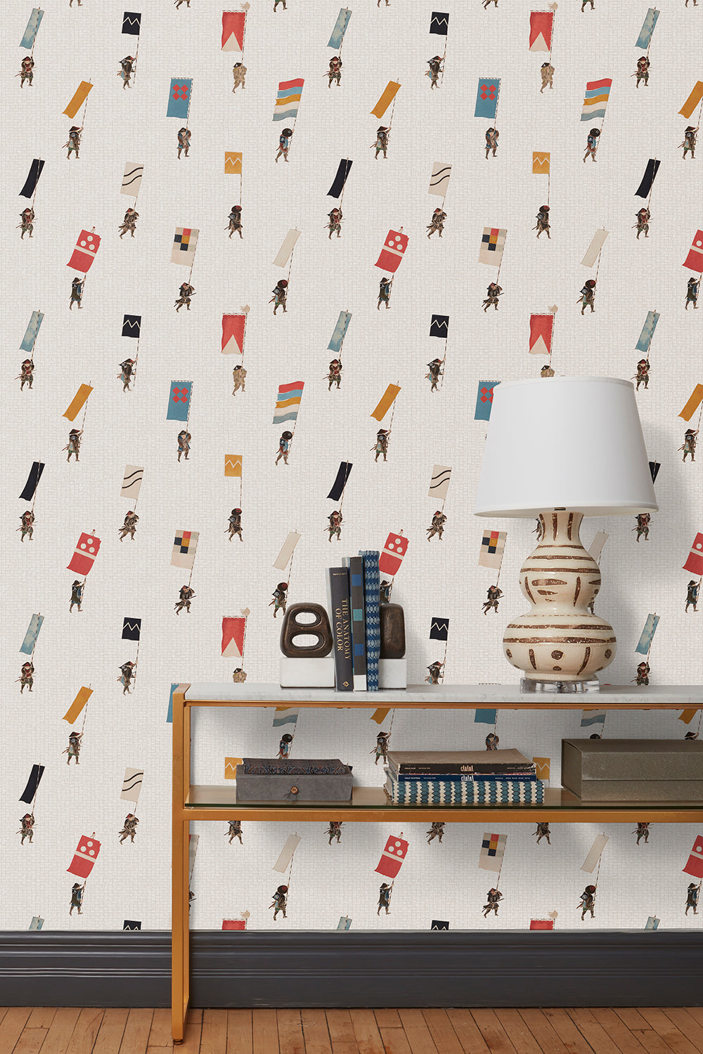 A table with books and a lamp in front of a wall papered in a pattern of Japanese soldiers carrying flags on a cream background.
