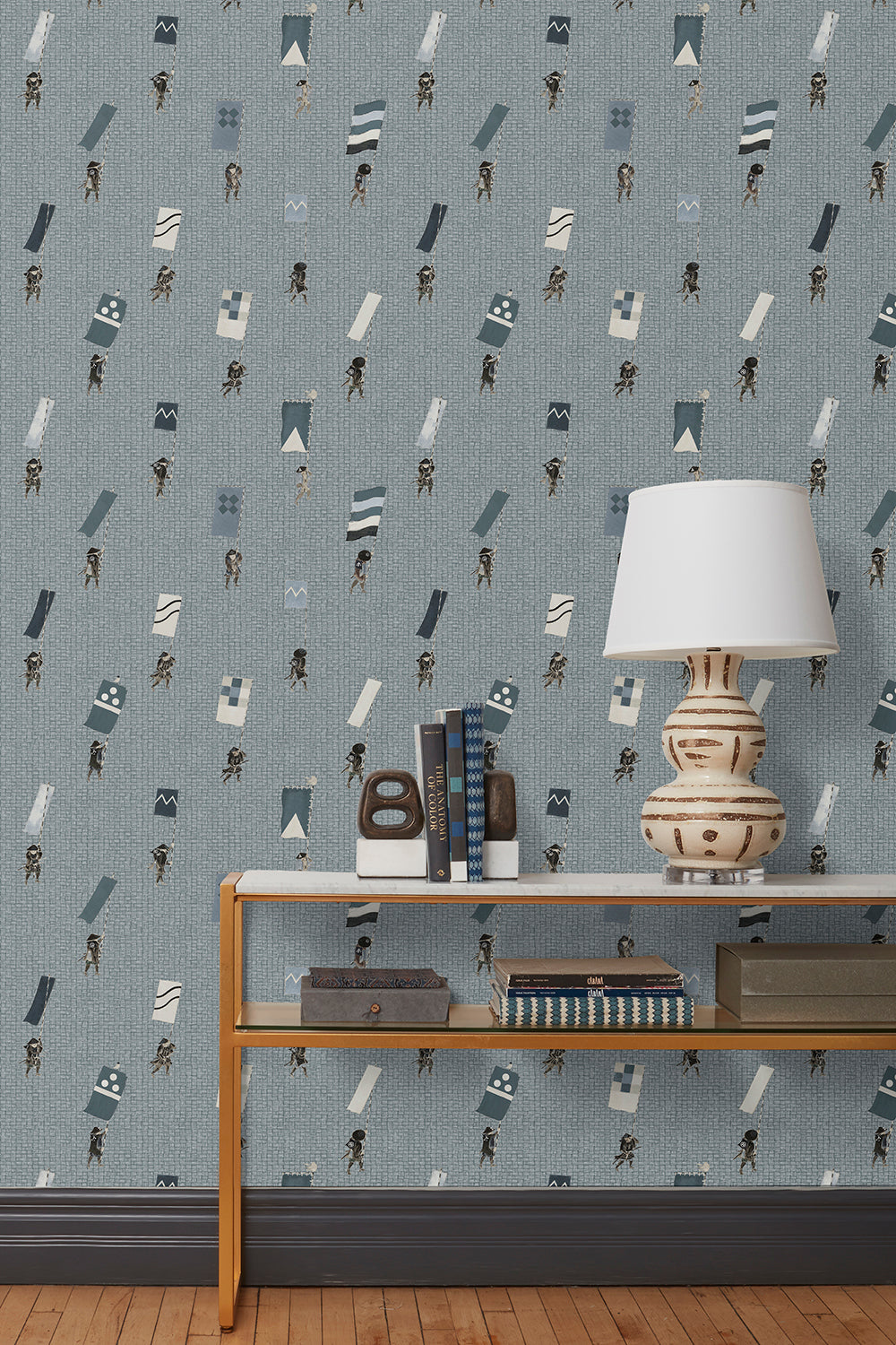 A table with books and a lamp in front of a wall papered in a pattern of Japanese soldiers carrying flags on a blue-gray background.