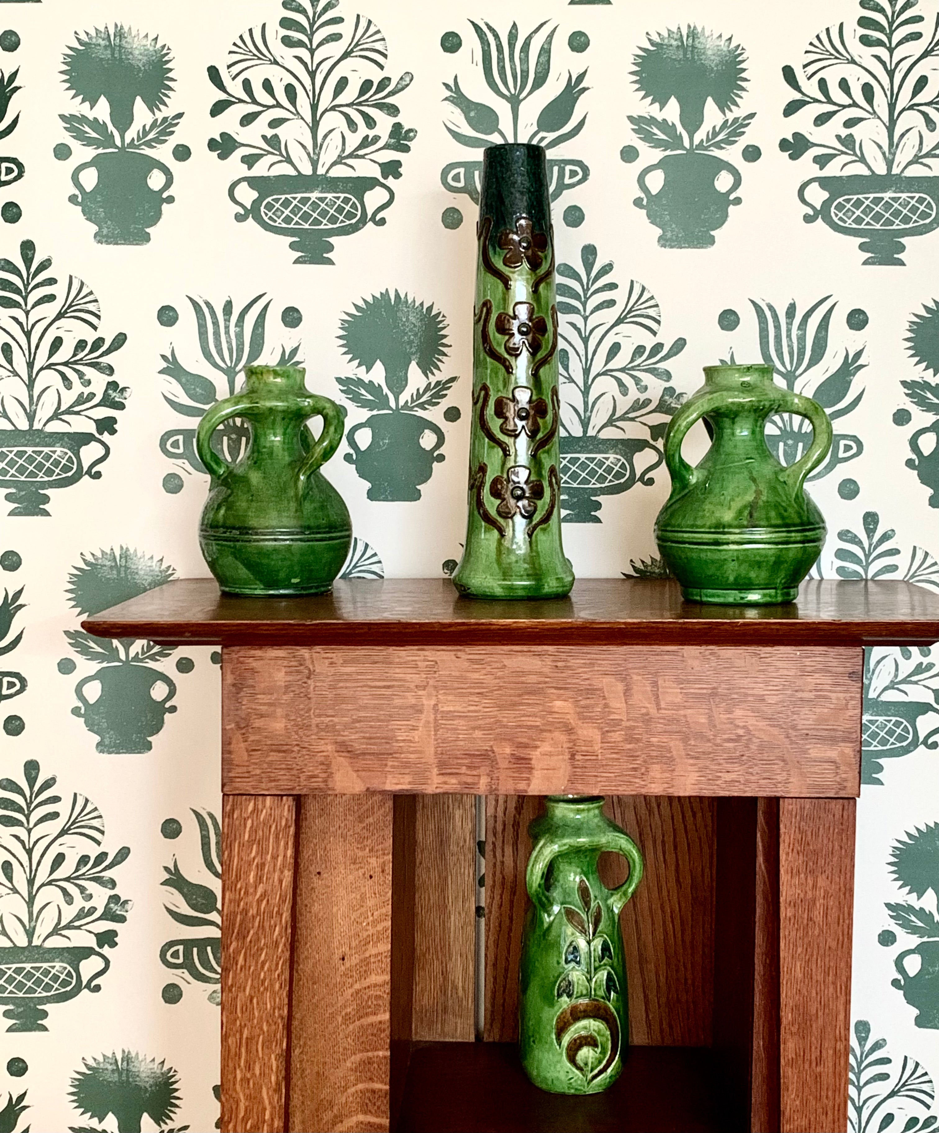 An end table and vases stand in front of a wall papered in a vase and plant print in green on a white field.