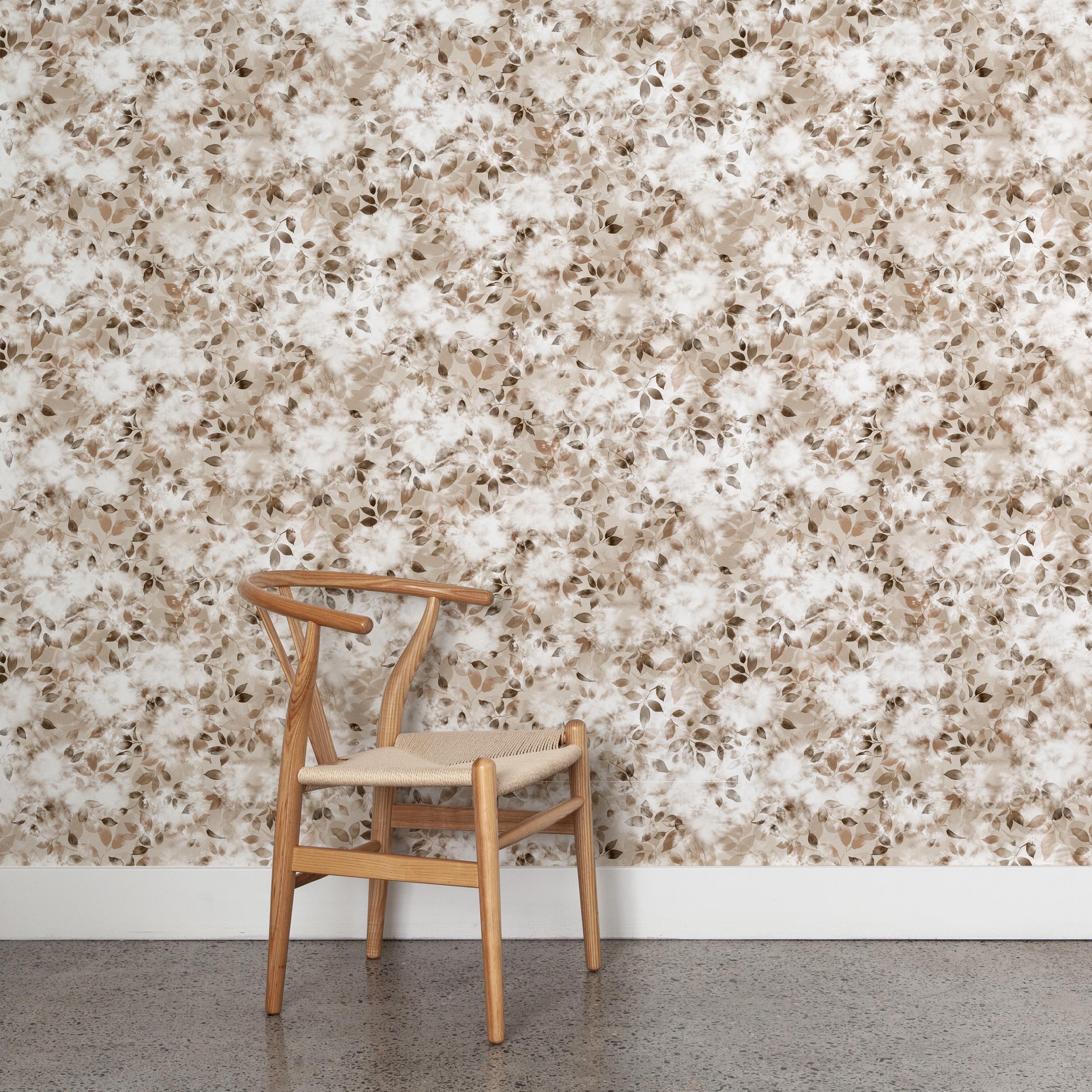 A wooden chair stands in front of a wall papered in a leaf print in shades of cream and tan with a mottled white overlay.