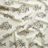 Close up of gently draped printed linen with a pattern of hand-drawn fish in black with tan and brown scales and scattered bubble dots on white. 
