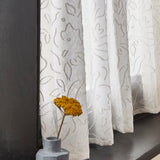 A window sill with a vase and wildflower against a sheer curtain in a hand drawn floral pattern in ivory.
