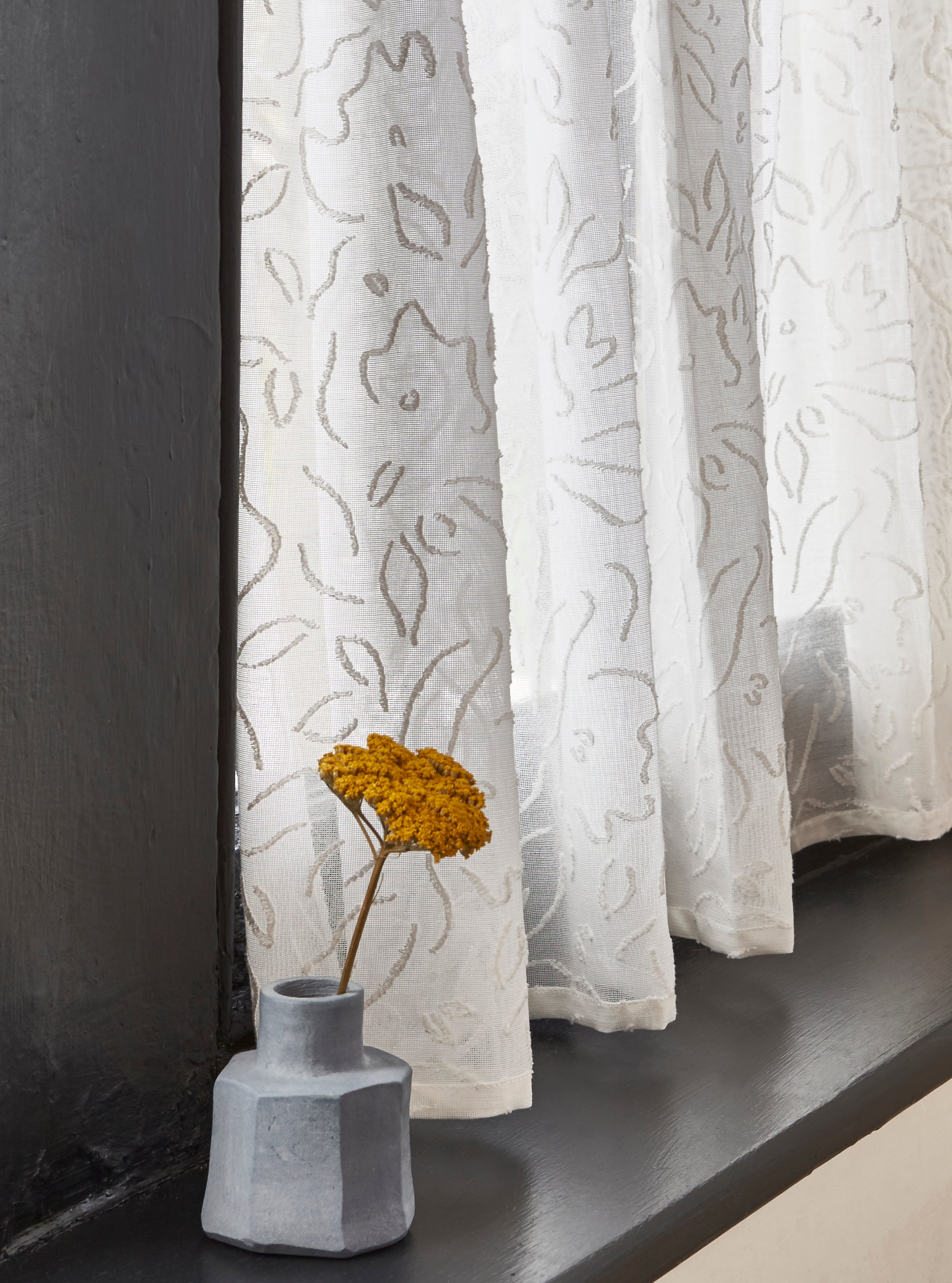 A window sill with a vase and wildflower against a sheer curtain in a hand drawn floral pattern in ivory.