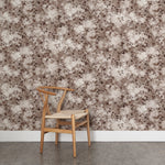 Angled wooden chair with woven seat on concrete floor against wallpaper in a painterly leaf print in shades of warm wine purple with a mottled white overlay.